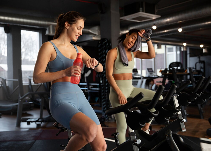 Две девушке едут на спортивном велосипеде одна смотрит на часы, другая вытирает пот со лба